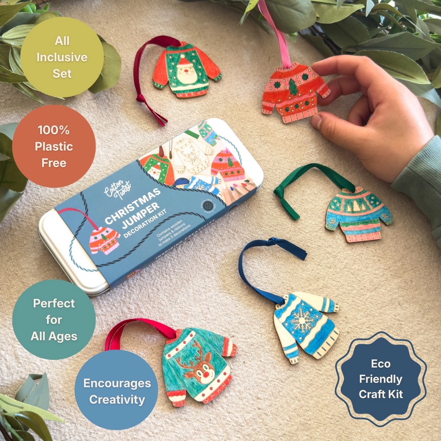 Christmas jumper craft kit shown with its tin packaging and several small sweater-shaped ornaments laid out on a beige surface, with a hand holding one ornament. Text callouts say it is an all inclusive set, 100% plastic free, perfect for all ages, encourages creativity, and is eco friendly.