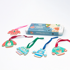 A Christmas jumper ornament kit with five colorful wooden sweater ornaments in different holiday patterns, hanging ribbons, and a blue metal tin box labeled 'Christmas Jumper' on a white background.