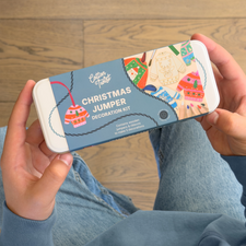 A person holding a tin of Cotton Twist Christmas Jumper Decoration Kit over a wooden floor and blue jeans.
