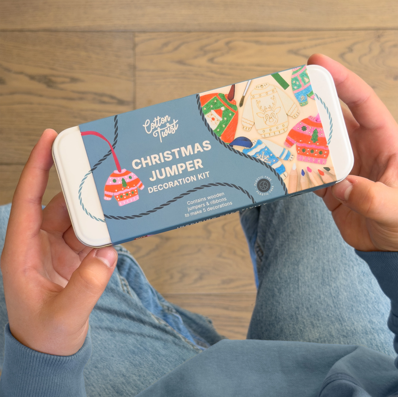 A person holding a tin of Cotton Twist Christmas Jumper Decoration Kit over a wooden floor and blue jeans.