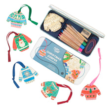 A Christmas jumper decoration kit shown with an open metal tin containing wooden ornaments, colored pencils, and a paintbrush, plus several completed colorful Christmas sweater ornaments arranged around it on a white background.