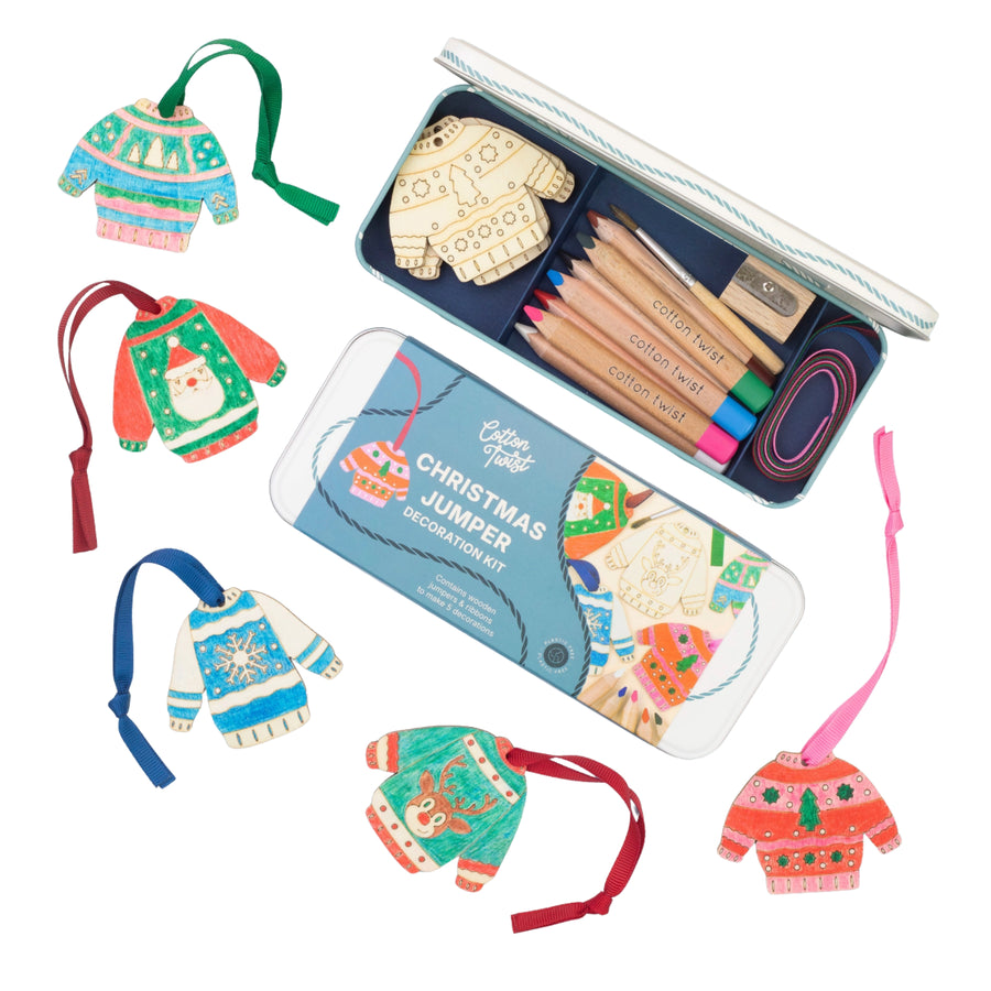 A Christmas jumper decoration kit shown with an open metal tin containing wooden ornaments, colored pencils, and a paintbrush, plus several completed colorful Christmas sweater ornaments arranged around it on a white background.